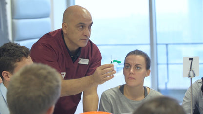 Teacher in maroon shirt pointing at a whiteboard with students in a classroom setting VeinTrain flat-pack vein simulator for phlebotomy and cannulation training

